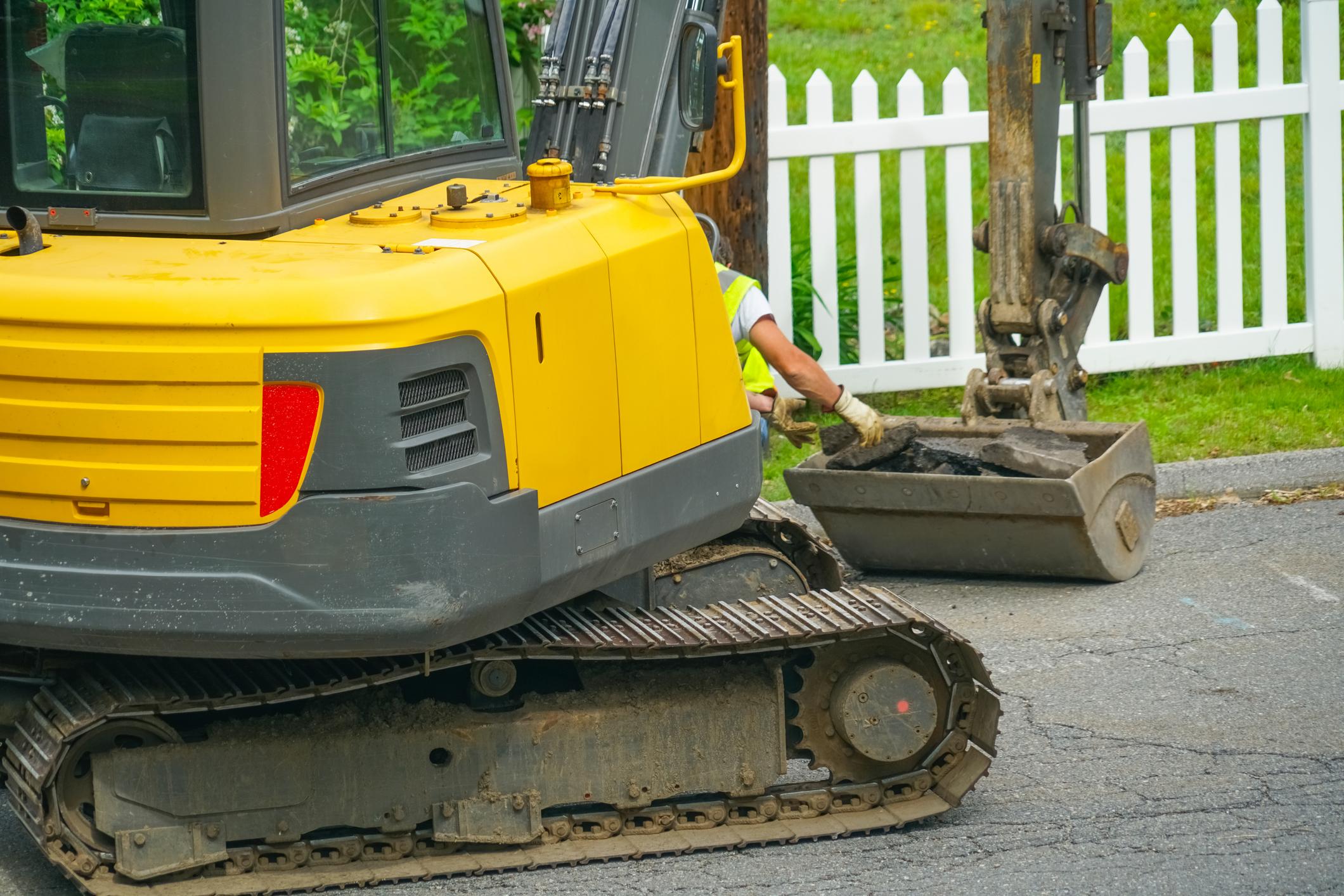 The image shows a yellow construction excavator in motion, preparing to dig on a residential street.
AI-generated content may be incorrect.