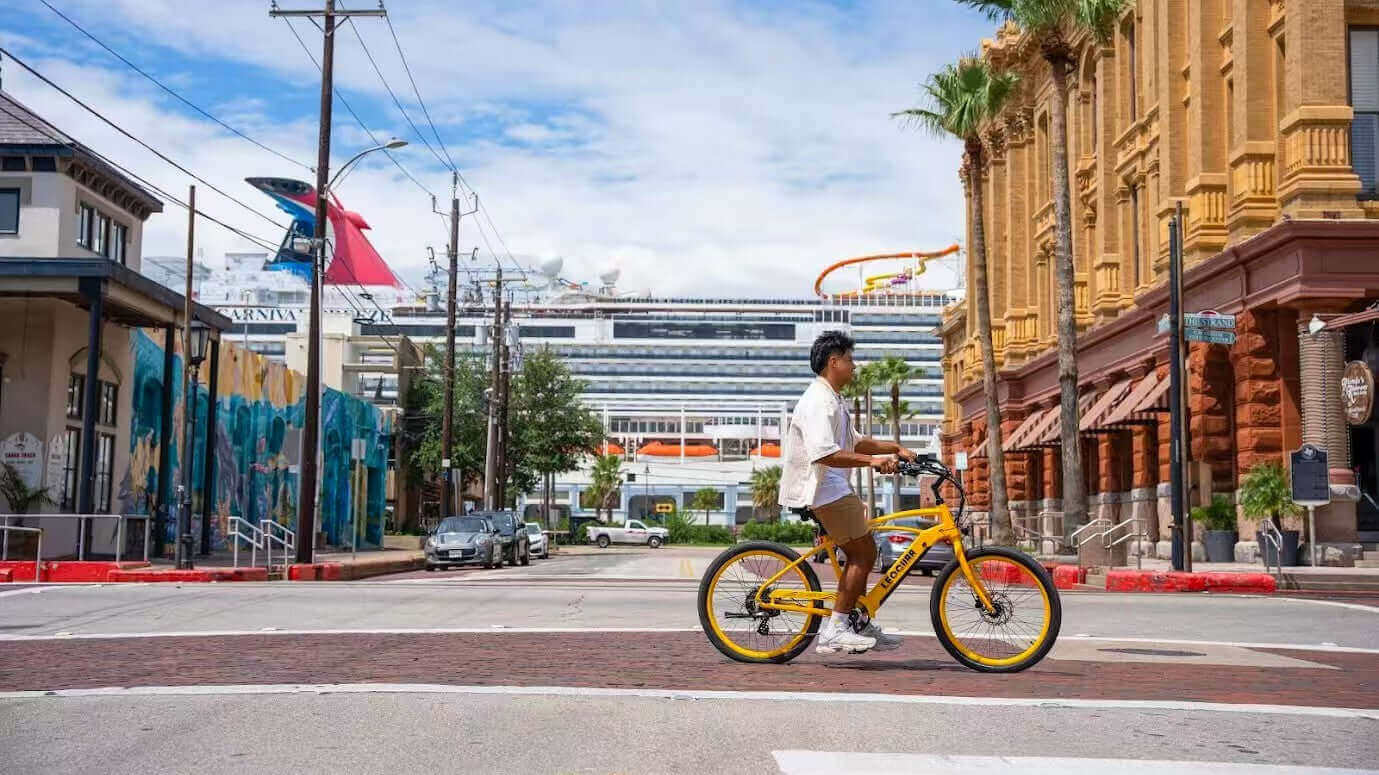 A person rides a yellow bicycle down a street with palm trees, a modern building, and a carnival float in the background.
AI-generated content may be incorrect.
