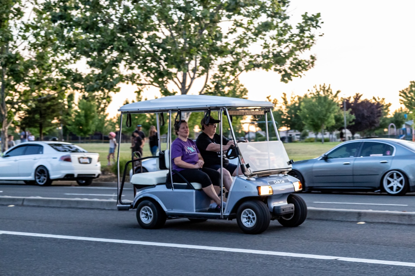 The image depicts a two-seater golf cart being driven by two people along a road with parked cars, under a clear, leafy sky.
AI-generated content may be incorrect.