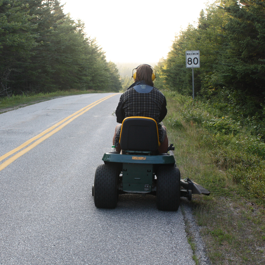 A person is riding a green push mower down a rural road with a sign indicating a speed limit of 80 km/h.
AI-generated content may be incorrect.