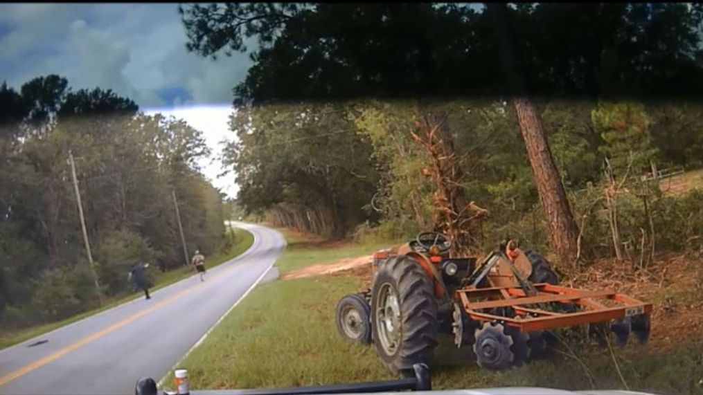 The image shows a rural scene with a large, orange tractor towing a trailer, navigating a winding forest road under a cloudy sky.
AI-generated content may be incorrect.