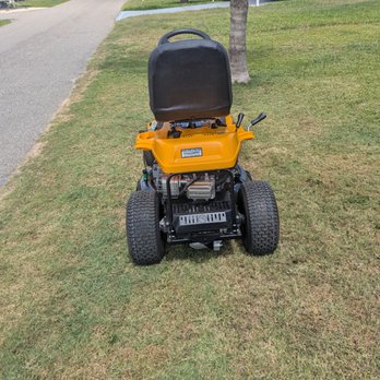 The image shows a yellow, battery-powered lawn mower sitting on a grassy lawn, with a side view, and some grass and a road visible in the background.
AI-generated content may be incorrect.